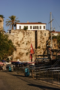 A historic stone building with red-tiled roof sits atop an old stone wall under a clear blue sky. Tall palm trees flank the structure. Below the wall, a marina is visible with moored ships and a Turkish flag. People can be seen walking along the cobblestone path, with market stalls and cafes nearby.