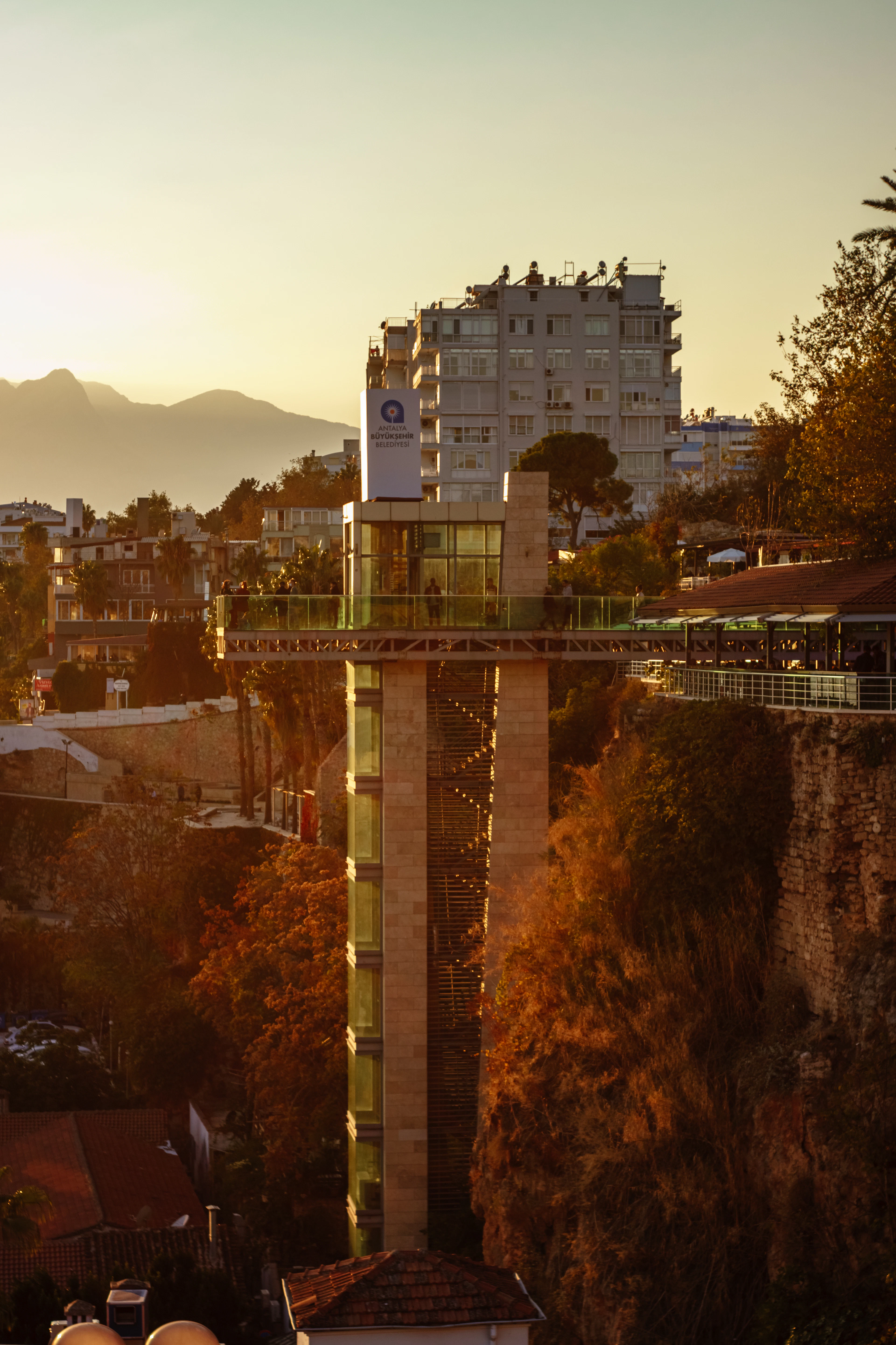 The glass panoramic lift rising up the cliff face below Antalya's old town at sunset, with the clifftop terraces and apartment blocks lit gold behind it.