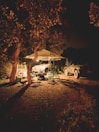 A nighttime camping scene with two vehicles parked under a canopy illuminated by string lights. The area is surrounded by trees, creating a cozy and secluded atmosphere. Various camping equipment and supplies are scattered around the site.