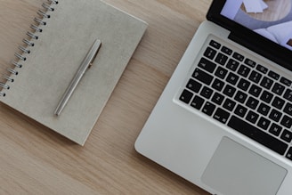 A silver laptop with a visible keyboard sits on a light wooden surface next to a closed spiral-bound notebook and a metal pen.