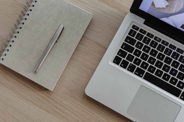 A silver laptop with a visible keyboard sits on a light wooden surface next to a closed spiral-bound notebook and a metal pen.