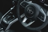 A close-up view of a car's interior focusing on the steering wheel and center console. The steering wheel features a prominent Toyota logo in the center, with modern design elements and control buttons. The gear shift lever is visible next to the steering wheel.