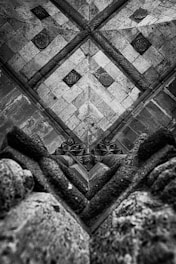 A geometric and symmetrical view of a ceiling, featuring intricate stonework with diagonal and diamond patterns. The design includes carved elements and decorative roundels, emphasizing gothic architectural details. The texture of the stone appears worn, adding to the historical and architectural depth.