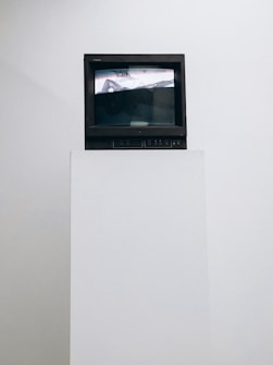 A vintage television set sits atop a white pedestal. The screen displays an abstract image in black and white with some distorted text visible. The surrounding background is plain white, emphasizing the contrast with the dark frame of the TV.