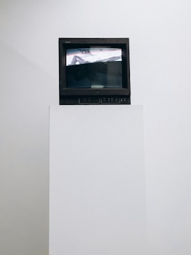A vintage television set sits atop a white pedestal. The screen displays an abstract image in black and white with some distorted text visible. The surrounding background is plain white, emphasizing the contrast with the dark frame of the TV.