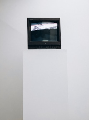 A vintage television set sits atop a white pedestal. The screen displays an abstract image in black and white with some distorted text visible. The surrounding background is plain white, emphasizing the contrast with the dark frame of the TV.