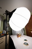 A photography setup with a large softbox light on a stand illuminating a table. On the table, there are various objects including jars, plates with cookies, and decorative items. The background includes a brick wall and part of a kitchen countertop with miscellaneous items.