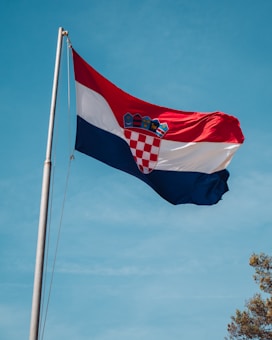 A Croatian flag waving on a tall flagpole against a clear blue sky. The flag features a red, white, and blue tricolor with the national coat of arms in the center. A few tree branches are visible in the lower right corner.