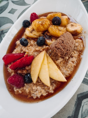A dish featuring a bowl of creamy oatmeal topped with caramelized banana slices, fresh blueberries, raspberries, and sliced pears. The oatmeal is garnished with a scoop of brown sugar or nut butter, and is surrounded by a pool of syrup or sauce.