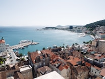 A coastal cityscape featuring a harbor with several boats and yachts docked. The city is characterized by its historic buildings with red-tiled roofs, a mix of modern and old architecture. In the background, the sea stretches into the horizon with hills on the distant shore.