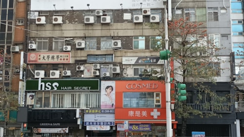 A street view of a multi-story building featuring numerous air conditioning units installed on its facade. The ground floor consists of various shops with signs in different languages, including a hair salon and a pharmacy. There are trees lining the street, some traffic lights, and various advertisements.