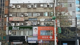 A street view of a multi-story building featuring numerous air conditioning units installed on its facade. The ground floor consists of various shops with signs in different languages, including a hair salon and a pharmacy. There are trees lining the street, some traffic lights, and various advertisements.