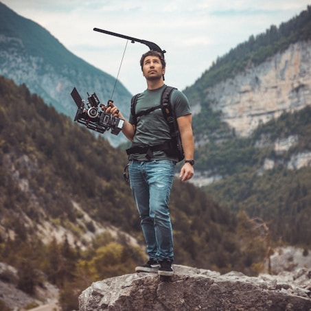 A man stands on a rock, holding a professional camera rig. He wears casual clothing and seems to be in a mountainous landscape with greenery in the background. The scene conveys a sense of adventure and filmmaking in nature.