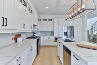 A modern kitchen with white cabinetry and stainless steel appliances. The countertops are light-colored and there is a large central island with a sink. The space is illuminated by a rectangular light fixture with exposed bulbs. A wooden cutting board and a small plant add decorative touches.
