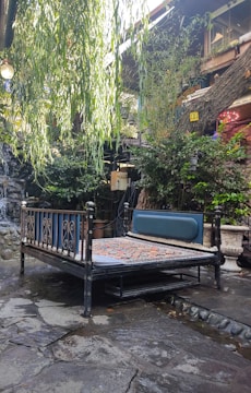 An outdoor setting featuring a vintage metal bed frame with a colorful patchwork quilt. The area surrounding the bed is lush with greenery, including overhanging willow branches and various potted plants. The background includes a stone path and a rustic building exterior with wooden textures.