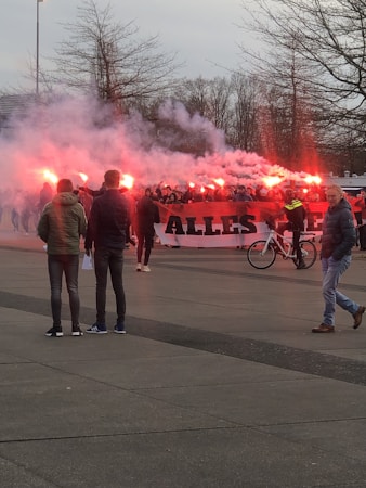 A group of people are gathered holding red flares, creating smoke. They stand behind a large banner with partially visible text. The scene takes place outdoors on a paved area with leafless trees in the background. Some people are walking nearby, and one person is riding a bicycle.