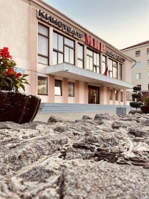 A building with a sign in Cyrillic script, possibly a cinema, features large windows and an entrance with a canopy. The foreground includes a stone pathway and a planter with red flowers. The sky is clear, and adjacent buildings are visible.