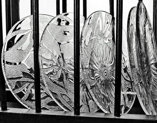 Decorative glass plates featuring intricate floral and geometric patterns are arranged in a row behind vertical black bars, with strong contrasts creating a prominent texture.