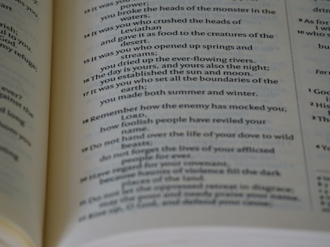 A close-up view of an open book, revealing text passages with poetic and religious themes. The page features numbered verses and italicized text, suggesting it might be a page from a Bible or other religious text.