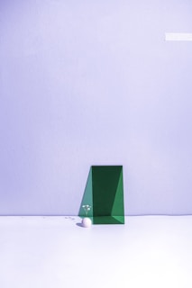 A minimalist composition featuring a green geometric shape positioned against a pale lavender wall. A small white sphere sits on the floor next to the shape, adorned with delicate white flowers, creating a subtle and balanced aesthetic.
