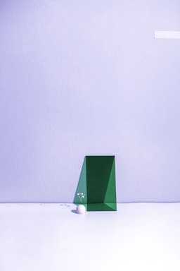 A minimalist composition featuring a green geometric shape positioned against a pale lavender wall. A small white sphere sits on the floor next to the shape, adorned with delicate white flowers, creating a subtle and balanced aesthetic.