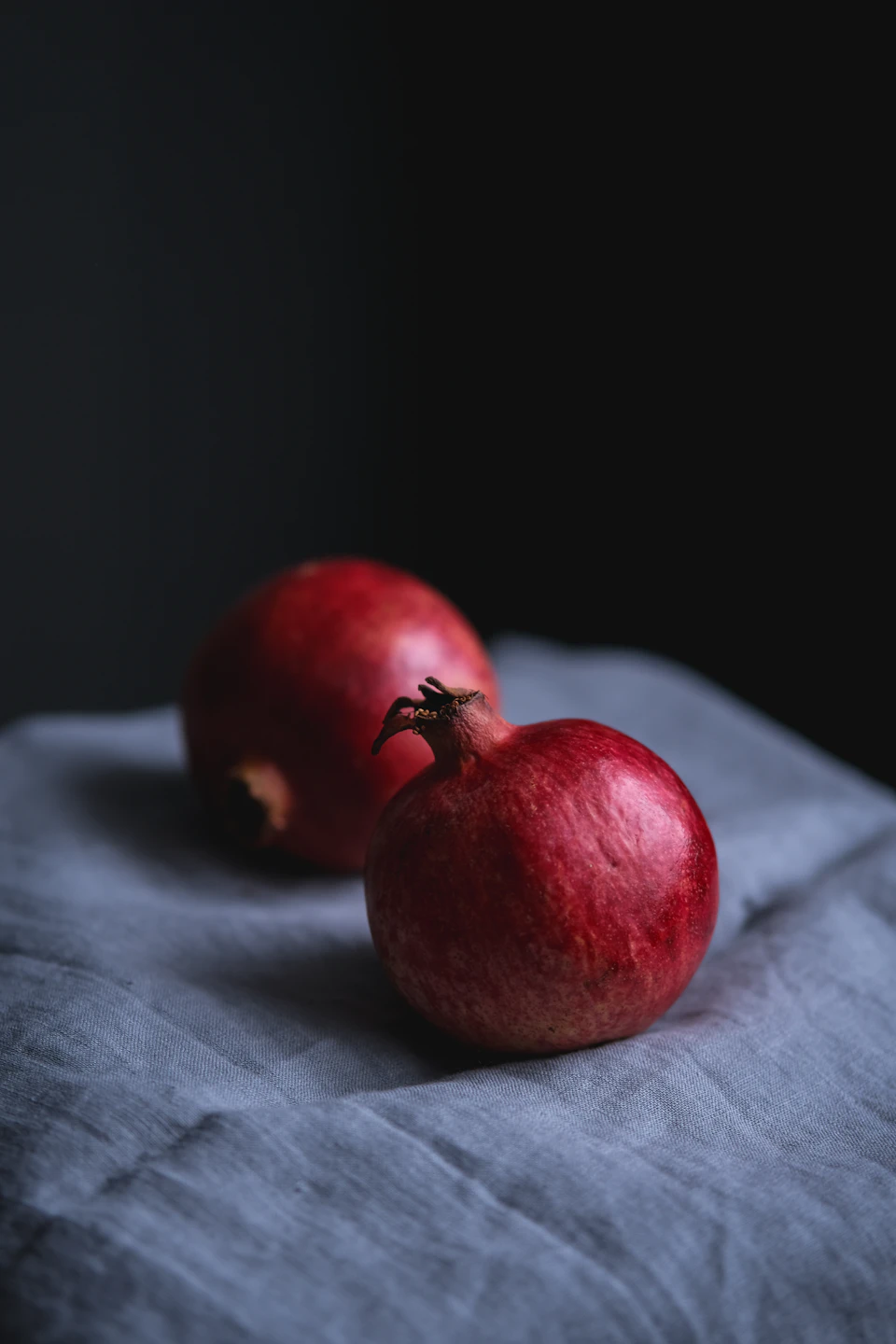Two red pomegranates rest on a gray cloth against a black background together
