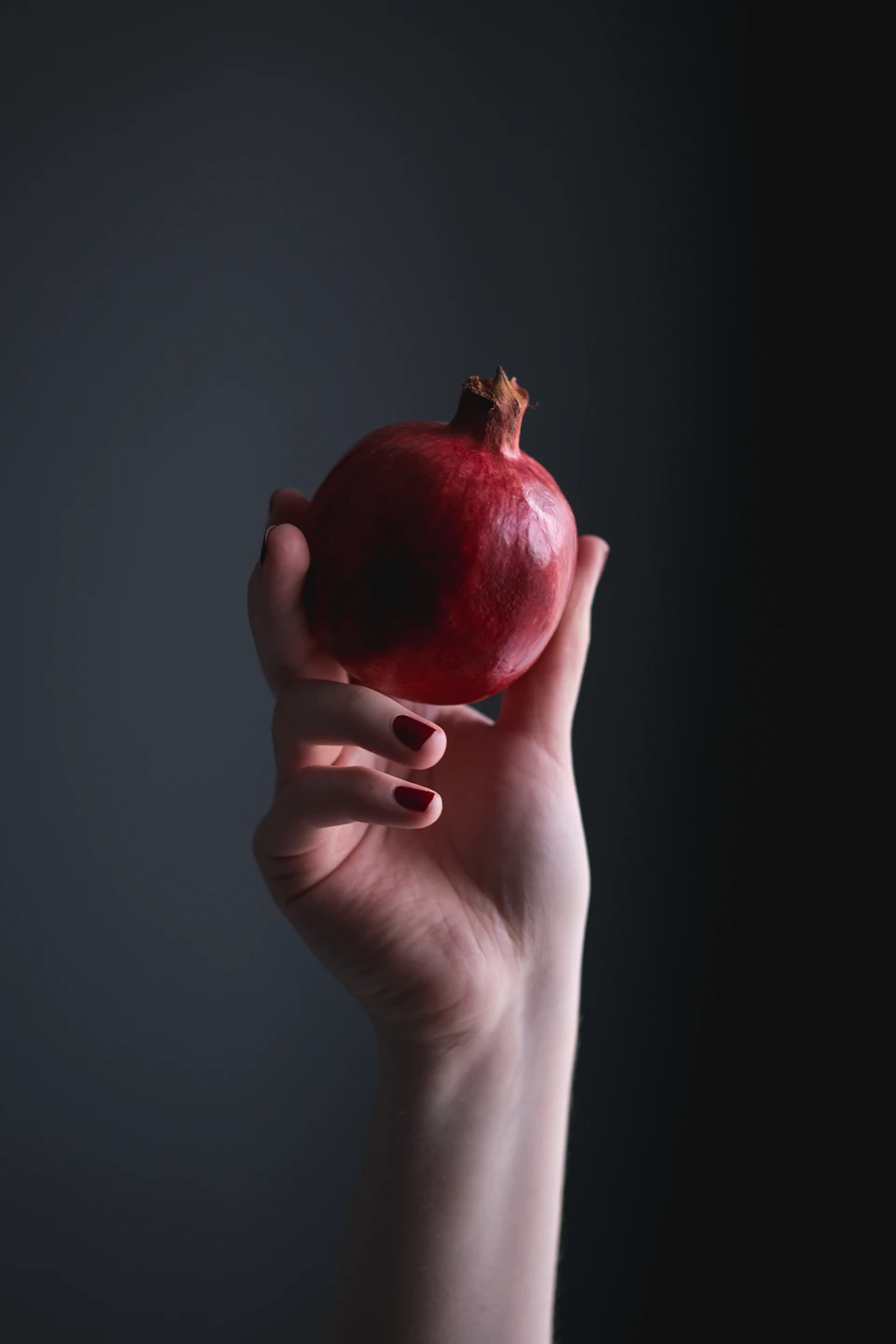 The image shows a hand holding up a single, vibrant red pomegranate against a stark black background. The pomegranate is positioned centrally within the frame and appears to be the main focus of the image.
