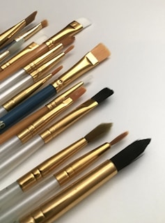 A collection of artist paintbrushes with varying sizes and shapes laid out on a white surface. The brushes have golden ferrules and handles in shades of silver, wood, and blue.