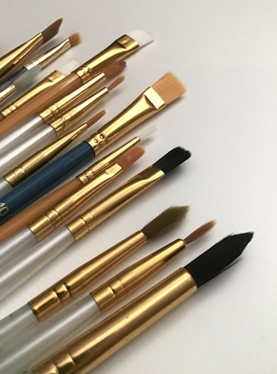 A collection of artist paintbrushes with varying sizes and shapes laid out on a white surface. The brushes have golden ferrules and handles in shades of silver, wood, and blue.