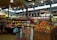 A busy market scene with a variety of fresh produce including onions and potatoes, packaged snacks, and grocery items displayed on wooden and metal shelving. Bright overhead lighting illuminates the spacious interior with a high ceiling, where shoppers are seen browsing. Visible signage indicates pricing for different items.