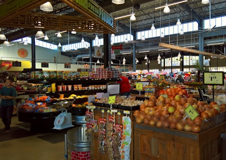 A busy market scene with a variety of fresh produce including onions and potatoes, packaged snacks, and grocery items displayed on wooden and metal shelving. Bright overhead lighting illuminates the spacious interior with a high ceiling, where shoppers are seen browsing. Visible signage indicates pricing for different items.