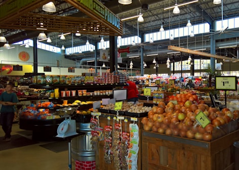 A busy market scene with a variety of fresh produce including onions and potatoes, packaged snacks, and grocery items displayed on wooden and metal shelving. Bright overhead lighting illuminates the spacious interior with a high ceiling, where shoppers are seen browsing. Visible signage indicates pricing for different items.