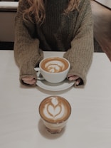 A person wearing a cozy knit sweater holds a large cup of coffee with latte art. In the foreground, another cup of coffee with similar latte art sits on a white table.