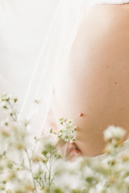 A close-up of a pregnant belly surrounded by delicate, small white flowers. The soft focus on the flowers in the foreground and the gentle lighting create a peaceful and dreamy atmosphere.
