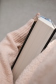 A person wearing a cozy, light-colored sweater is holding a closed hardcover book. The focus is on the hand and book, suggesting a calm and relaxed setting.