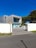 A modern, minimalist house with a rectangular design and large glass windows. The building has a combination of wooden and concrete textures, surrounded by a clean courtyard with sparse greenery. The sky is bright and clear with a rich blue color.