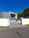 A modern, minimalist house with a rectangular design and large glass windows. The building has a combination of wooden and concrete textures, surrounded by a clean courtyard with sparse greenery. The sky is bright and clear with a rich blue color.