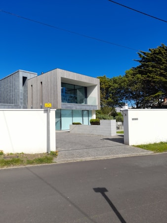 A modern, minimalist house with a rectangular design and large glass windows. The building has a combination of wooden and concrete textures, surrounded by a clean courtyard with sparse greenery. The sky is bright and clear with a rich blue color.