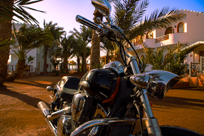 A shiny motorcycle is parked in front of a luxurious villa, surrounded by tall palm trees. The sunlight creates reflections on the chrome parts of the bike, and the setting gives a warm, tropical feel.