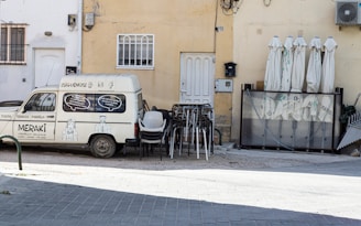 An old white van with artistic illustrations and text is parked against a pale yellow building. Next to the van, there are stacked chairs and folded umbrellas, suggesting an outdoor seating area. The wall features a few windows and an air conditioning unit.
