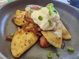 Two golden-brown, pan-fried dumplings are garnished with sautéed onions, a generous dollop of sour cream, and thinly sliced green onions, presented in a dark gray bowl.
