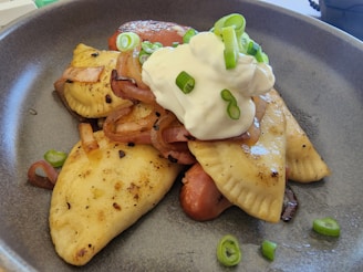 Two golden-brown, pan-fried dumplings are garnished with sautéed onions, a generous dollop of sour cream, and thinly sliced green onions, presented in a dark gray bowl.