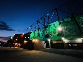 A building facade illuminated with green lights features large banners or murals showing historical sports scenes, likely related to soccer, with visible dates and trophies. The sky is evening blue with scattered clouds, and the ground is paved, partially lit by the building's exterior lights.