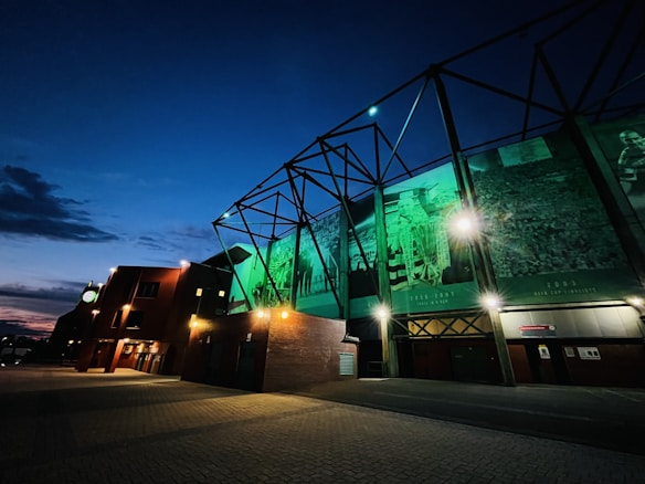 A building facade illuminated with green lights features large banners or murals showing historical sports scenes, likely related to soccer, with visible dates and trophies. The sky is evening blue with scattered clouds, and the ground is paved, partially lit by the building's exterior lights.