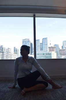 A person sitting indoors in a yoga pose next to a large window overlooks a cityscape with tall buildings. The lighting suggests it might be dawn or dusk, creating a silhouette of the person.