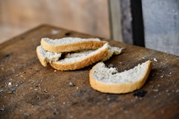 Several slices of white bread are scattered on a worn, wooden surface. The surroundings appear rustic, with the bread showing a simple, homemade quality.