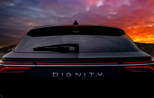 The back of a dark-colored SUV is prominently displayed with the word 'DIGNITY' on its rear. The sunset in the background casts orange, pink, and purple hues across the sky, creating a dramatic visual contrast with the vehicle.