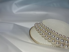 A close-up view of a string of lustrous pearls artistically arranged on top of a textured white surface, possibly a seashell, with a soft and smooth draped fabric as the background.