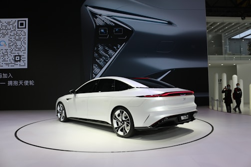 A sleek, white sedan is displayed on a showroom floor with a large digital backdrop featuring a close-up of the car's headlight design. The environment appears modern and minimalistic, and a QR code is visible on the left side of the image. Two people are casually observing the scene from a distance.