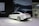 A sleek, white sedan is displayed on a showroom floor with a large digital backdrop featuring a close-up of the car's headlight design. The environment appears modern and minimalistic, and a QR code is visible on the left side of the image. Two people are casually observing the scene from a distance.
